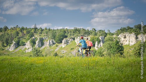 Zwei Personen fahren mit Fahrrädern auf einem grasbewachsenen Feld vor einer Felslandschaft.