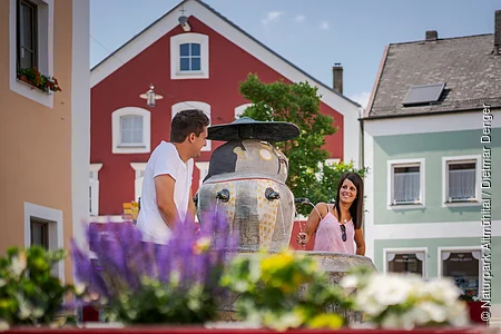 Zwei Personen stehen an einem Brunnen in einer bunten Stadtumgebung.