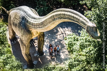 Große Dinosaurierstatue im Wald, Menschen stehen darunter und schauen nach oben.