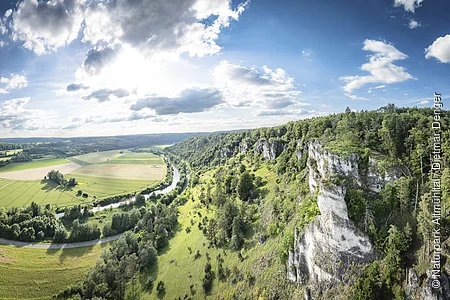 Landschaft mit grünen Feldern, einem Fluss und bewaldeten Hügeln unter einem bewölkten Himmel.