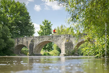 Steinbrücke mit drei Bögen über einen Fluss, umgeben von Bäumen und Vegetation.