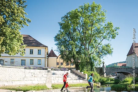 Zwei Personen überqueren auf Trittsteinen einen flachen Fluss, im Hintergrund Gebäude und Bäume.