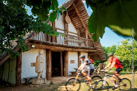 Holzhaus mit Statue davor und zwei Radfahrern, umgeben von Bäumen bei sonnigem Wetter