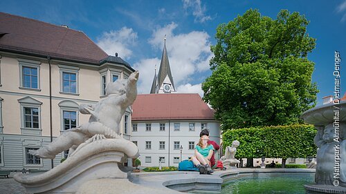 Paar sitzt am Rand eines Brunnens mit Steinfiguren vor historischen Gebäuden und einer Kirche bei blauem Himmel.