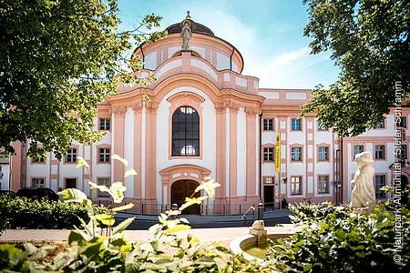Barockgebäude mit großer Fensterfront, Statue auf dem Dach und Springbrunnen im Vordergrund bei Sonnenschein