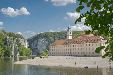 Flussufer mit Kiesstrand, altes Gebäude mit Turm und bewaldete Felsen unter blauem Himmel mit Wolken