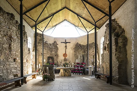 Innenraum einer Kapelle mit Altar, Kreuz, Kerzen und Blumen, umgeben von alten Steinmauern und Holzdecke