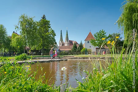 Park mit Teich, Wiese, Bäumen und Menschen vor historischen Gebäuden mit zwei Türmen bei blauem Himmel