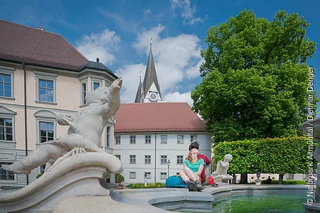 Paar sitzt am Rand eines Brunnens mit Steinfiguren vor historischen Gebäuden und einer Kirche bei blauem Himmel.