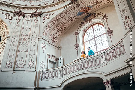 Zwei Personen stehen auf einem verzierten Balkon in einem historischen, mit Fresken geschmückten Innenraum.