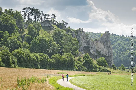 Zwei Radfahrer auf einem Weg vor bewaldeten Felsen mit einem Kreuz auf dem höchsten Felsen.
