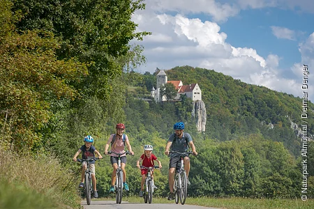Familie mit zwei Kindern fährt auf Fahrrädern auf einem Weg vor bewaldetem Hügel mit Burg im Hintergrund.