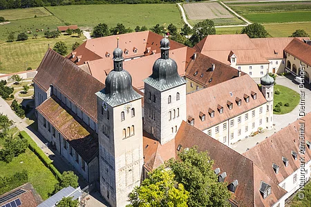 Luftaufnahme eines historischen Gebäudekomplexes mit zwei Kirchtürmen und roten Ziegeldächern in ländlicher Umgebung.