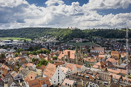 Luftaufnahme einer Stadt mit roten Dächern, Kirche mit zwei Türmen und bewaldetem Hügel im Hintergrund unter bewölktem Himmel.