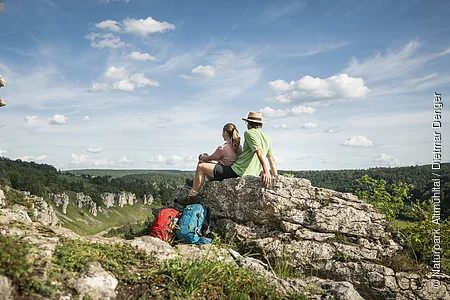 Zwei Wanderer sitzen auf einem Felsen und blicken auf bewaldete Hügel unter blauem Himmel mit Wolken.