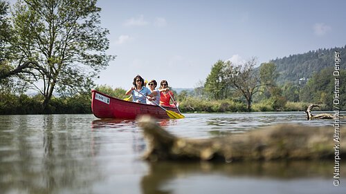 Bootwandern auf der Altmühl bei Kottingwörth Drei Personen paddeln in einem roten Kanu auf einem ruhigen Fluss mit Bäumen und Hügeln im Hintergrund.
