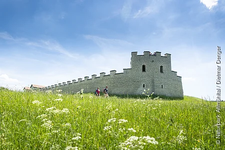 Zwei Wanderer gehen an einer historischen Steinmauer auf einer grünen Wiese unter blauem Himmel vorbei.