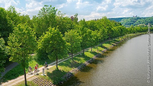 Fünf Radfahrer auf einem Weg entlang eines Flusses mit Bäumen und Hügeln im Hintergrund bei bewölktem Himmel.