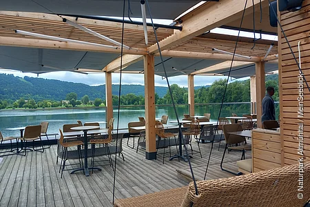 Überdachte Terrasse mit Tischen und Stühlen an einem See, im Hintergrund Bäume und bewaldete Hügel.