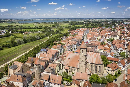 Luftaufnahme einer Stadt mit roten Dächern, zwei Türmen und einer großen Kirche, im Hintergrund grüne Felder und ein See.