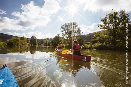 Zwei Personen paddeln in einem roten Kanu auf einem ruhigen Fluss bei sonnigem Himmel mit Wolken.