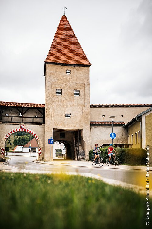 Zwei Radfahrer fahren an einem historischen Stadttor mit rotem Dach und Durchfahrt vorbei.