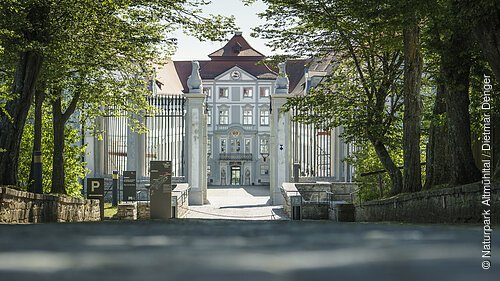 Eingangstor zu einem historischen Gebäude mit Bäumen und Schildern am Wegesrand bei Tageslicht.