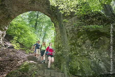 Vier Wanderer gehen einen Waldweg mit Steintreppe unter einem natürlichen Felsbogen hinab.