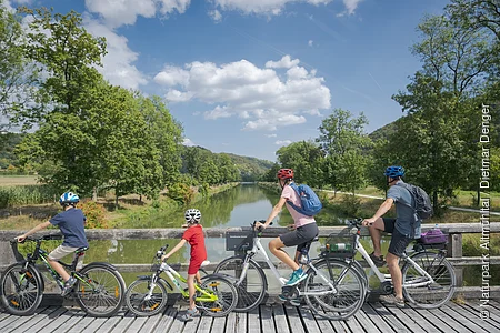 Vier Personen mit Fahrrädern auf einer Holzbrücke mit Blick auf einen Fluss und grüne Landschaft.