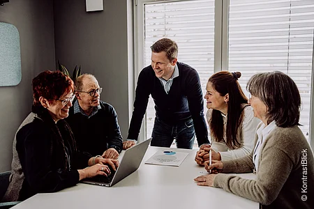 Fünf Personen sitzen und stehen an einem Tisch, eine Frau arbeitet an einem Laptop, alle wirken im Gespräch vertieft.