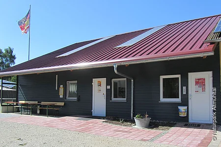 Eingang eines Gebäudes mit rotem Metalldach, zwei weißen Türen, Fenstern und einer Fahne im Hintergrund.