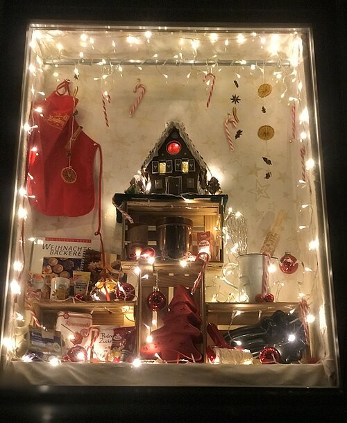 Weihnachtlich dekoriertes Fenster mit Lichterkette, Lebkuchenhaus, Backutensilien und roten Weihnachtskugeln.