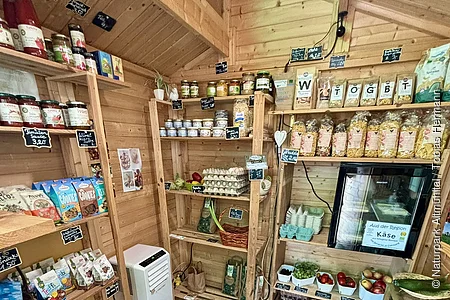 Kleiner Holzladen mit Regalen voller Lebensmittel, Eiern, Obst, Gemüse und Getränken.