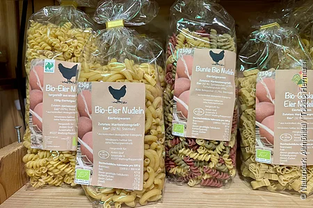 Verschiedene Packungen Bio-Eiernudeln in Plastikbeuteln auf Holzregal, Sorten und Formen sichtbar.