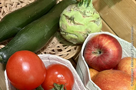 Tomaten, Äpfel, Gurken und Kohlrabi liegen auf einem Holztisch in Behältern und einem Korb.