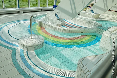 Innenbereich eines Schwimmbads mit kleinem Becken und bunten Fliesen in Regenbogenfarben am Boden.