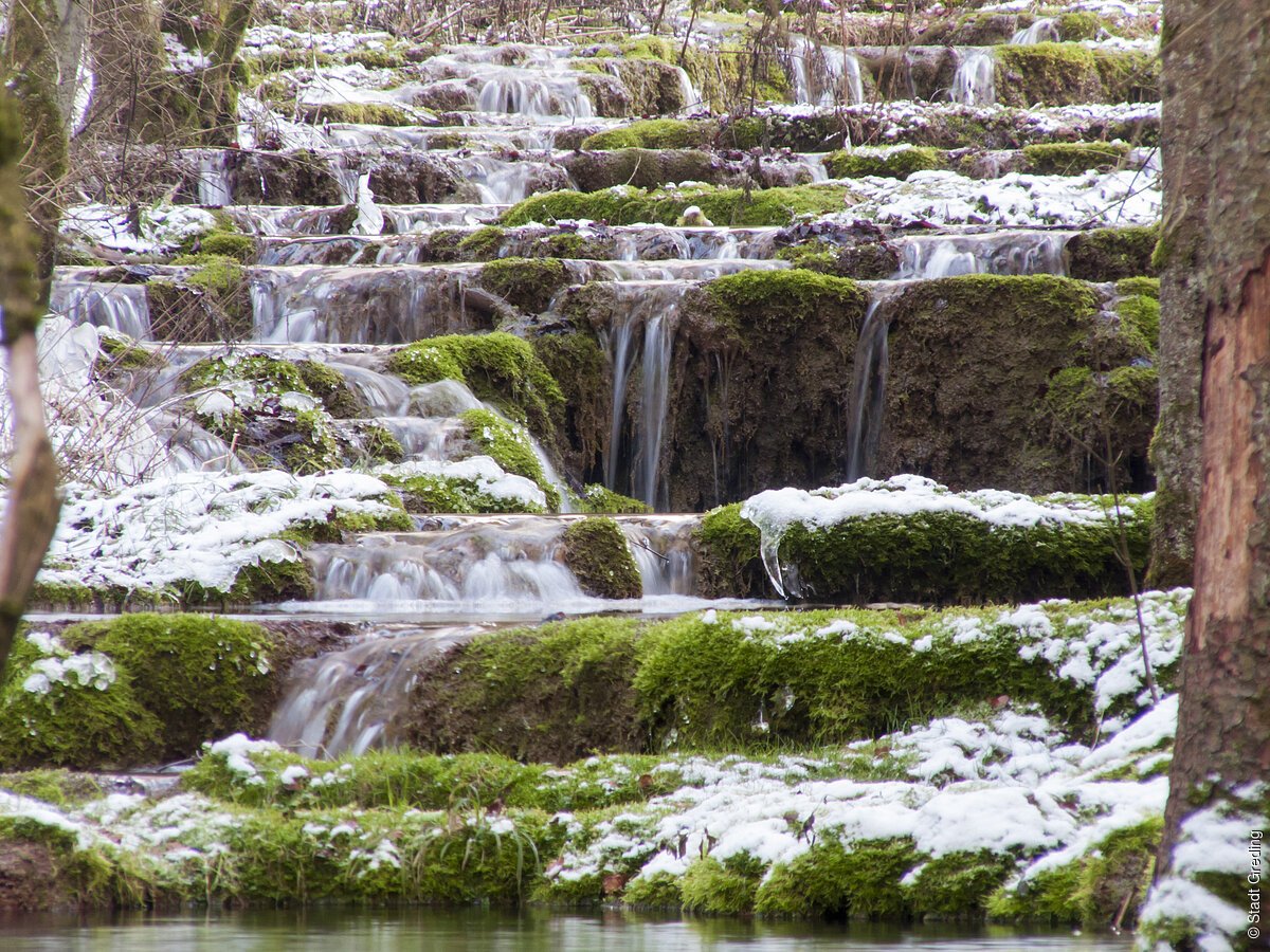 kleine Wasserfälle fließen über moosbedeckte Steinstufen mit leichtem Schneebelag im Wald