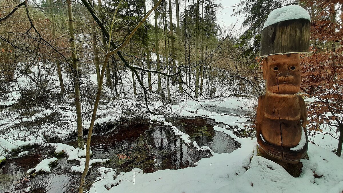 Holzfigur mit Schneehut neben einem kleinen Bach im verschneiten Wald im Winter.