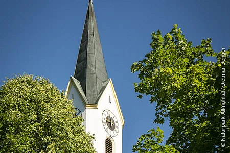 Kirchturm mit Uhr und Kreuz, umgeben von grünen Bäumen, vor blauem Himmel.