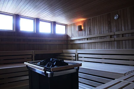 Innenraum einer Sauna mit Holzbänken, Holzverkleidung und einem Ofen mit schwarzen Steinen