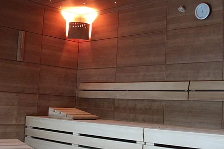 Innenraum einer Sauna mit hellen Holzbänken, einer Wandlampe und einer Uhr an der Wand