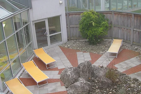 Innenhof mit drei gelben Liegestühlen, großen Steinen und einem Baum, umgeben von Glasfenstern und Holzzaun.