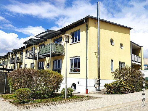 Mehrfamilienhaus mit gelber Fassade, Balkonen, runden Fenstern und Büschen bei sonnigem Wetter.
