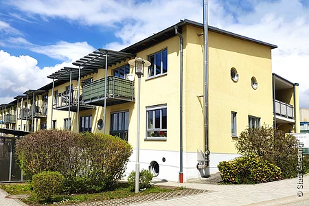 Mehrfamilienhaus mit gelber Fassade, Balkonen, runden Fenstern und Büschen bei sonnigem Wetter.