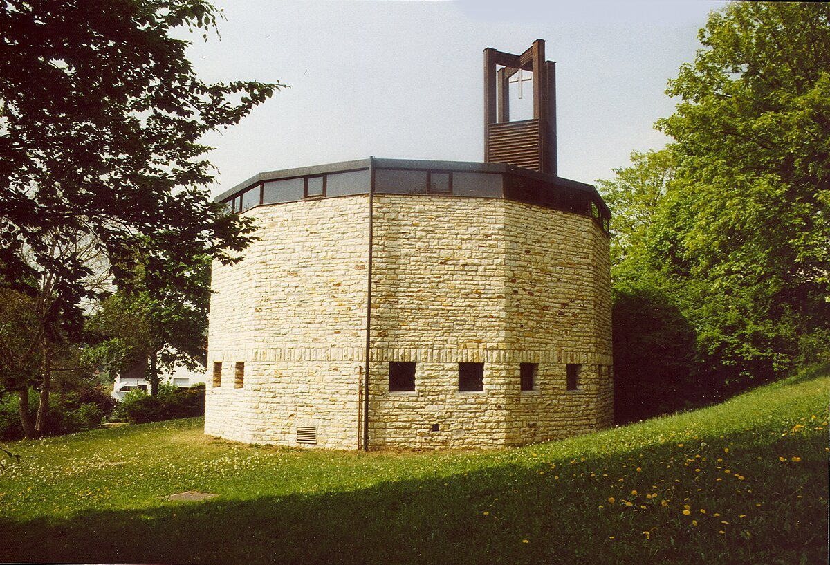 Runde Kirche aus hellem Stein mit kleinem Glockenturm und Kreuz, umgeben von Bäumen und Wiese.