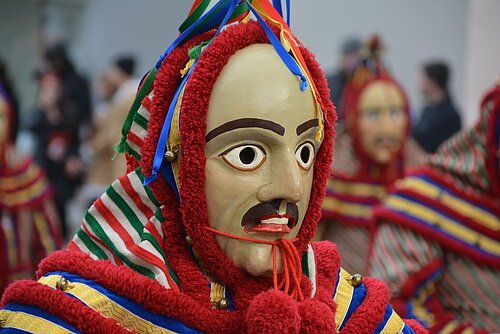 Nahaufnahme einer Person mit traditioneller bemalter Holzmaske und bunter Tracht bei einer Veranstaltung im Freien.