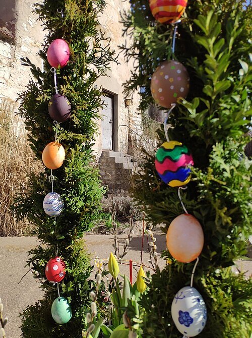 Zweige mit bunten Ostereiern hängen vor einer alten Steinmauer mit Tür und Treppe im Sonnenlicht.
