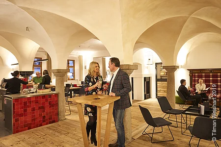 Zwei Personen stehen in gemütlichen Ambiente der Bar-Lounge der Post Berching und genießen am Stehtisch ein Getränk der Bar.