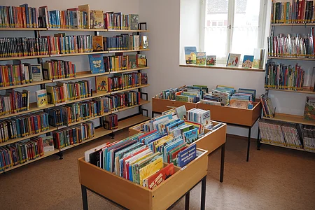 Kinderbücher in Holzregalen und auf Tischen in einem hellen Raum mit Fenster in einer Bibliothek.