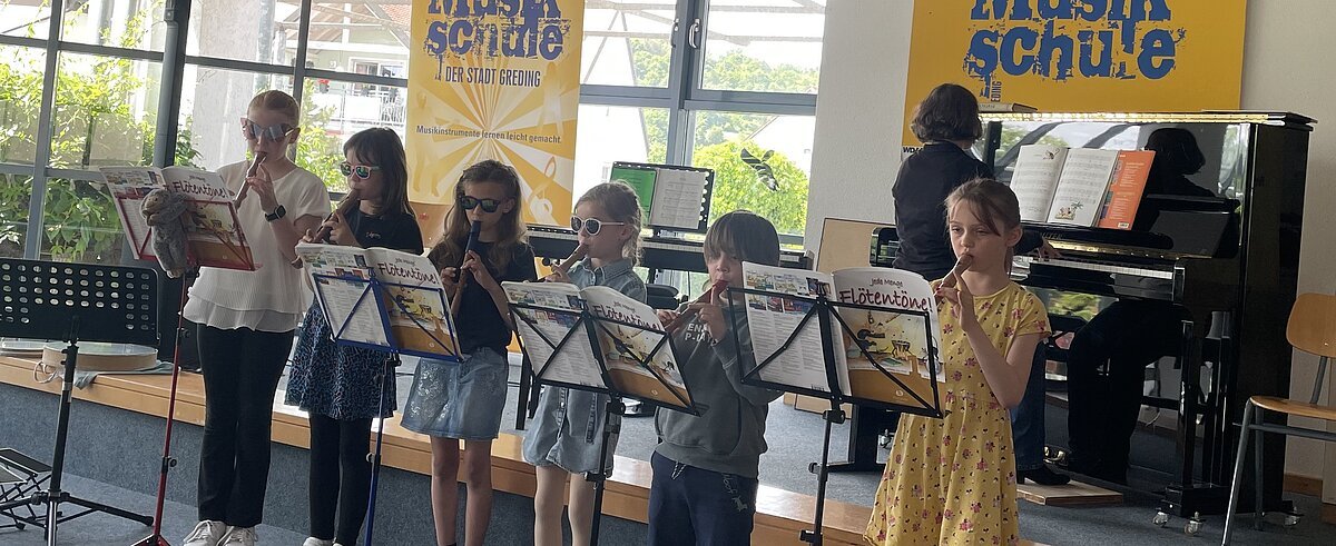 Musikschule Sechs Kinder spielen Blockflöte auf einer Bühne, eine Person spielt Klavier, im Hintergrund Banner der Musikschule.